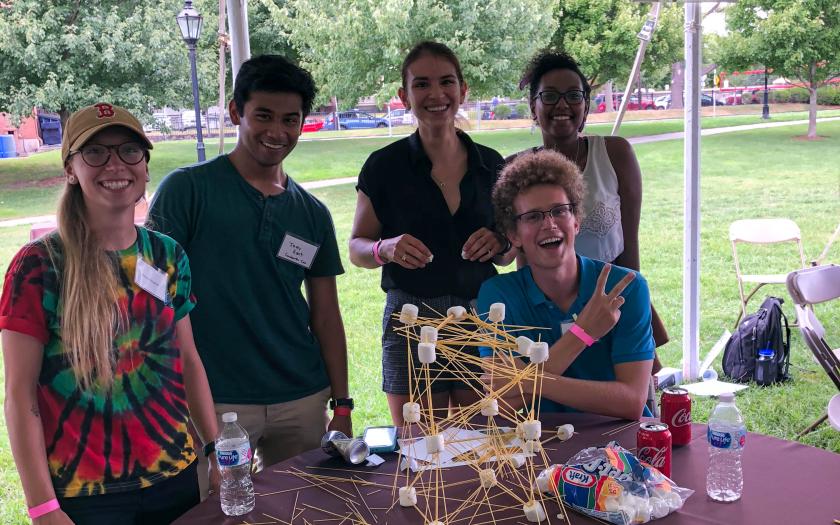 Students posing at an outdoor table with a structure built with spaghetti and marshmallows
