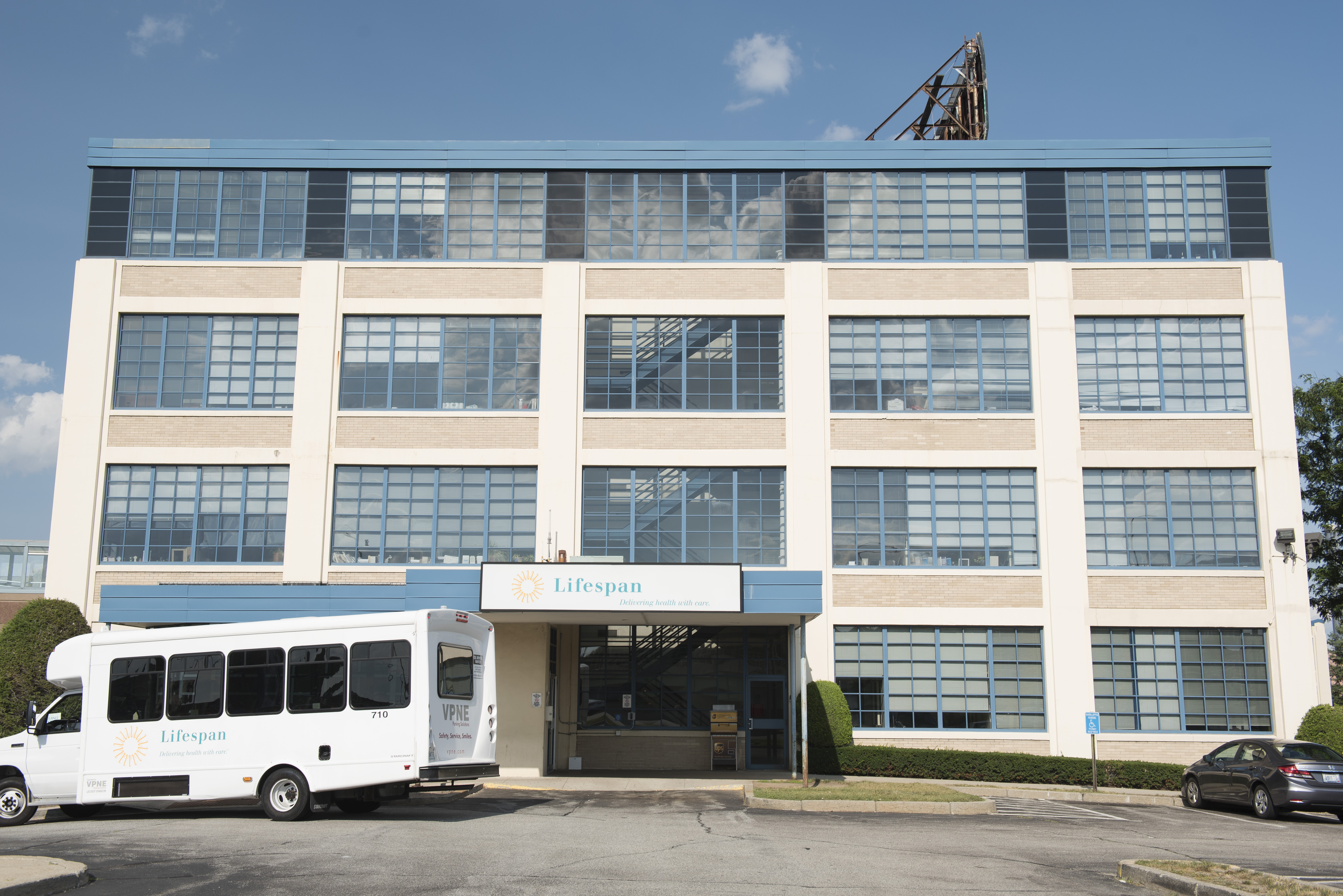 front view of a blue and grey modern building with a small white bus and parking in front